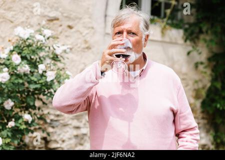 Homme barbu senior avec des cheveux gris buvant du vin rouge près du bâtiment pendant la fête de mariage contre des personnes sur fond flou Banque D'Images