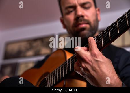 Un musicien masculin à barbe court de talent en turtleneck noir jouant du chant sur une guitare acoustique brune tout en étant assis près d'un mur carrelé dans la pièce Banque D'Images