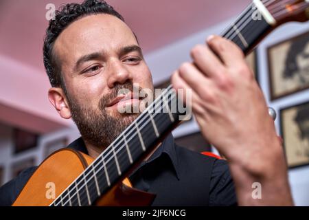 Musicien masculin barbu de talent en turtleneck noir jouant de la chanson sur guitare acoustique brune tout en étant assis près du mur de tuiles dans la pièce Banque D'Images