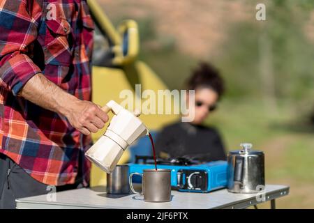 Vue latérale mâle anonyme touriste en chemise à carreaux remplissant une tasse de métal avec du café fraîchement infusé de la marmite de moka tout en prenant le petit déjeuner près de la camionnette pendant Banque D'Images