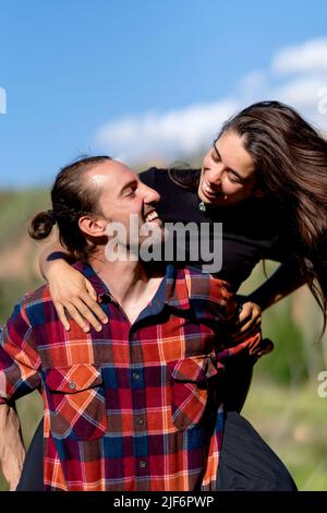 Homme gai en chemise à carreaux souriant et donnant le pigeyback à la petite amie avec de longs cheveux sombres tout en passant la journée de week-end d'été dans la campagne Banque D'Images