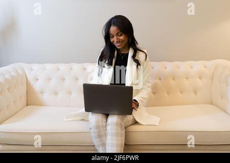 Femme entrepreneure souriante en costume assise sur un canapé et parcourant un netbook moderne tout en travaillant à distance dans le salon de la maison Banque D'Images