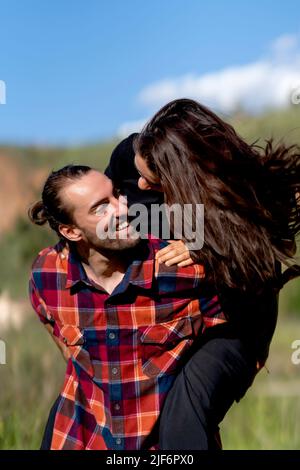 Homme gai en chemise à carreaux souriant et donnant le pigeyback à la petite amie avec de longs cheveux sombres tout en passant la journée de week-end d'été dans la campagne Banque D'Images