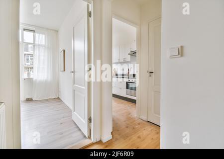 Couloir étroit avec murs et portes blancs menant à une chambre spacieuse avec fenêtres et parquet dans un appartement moderne Banque D'Images