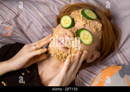 Une femme fait un masque cosmétique à la maison à partir de flocons d'avoine sur son visage et concombre.Traitements anti-âge, concept de soins de la peau. Banque D'Images