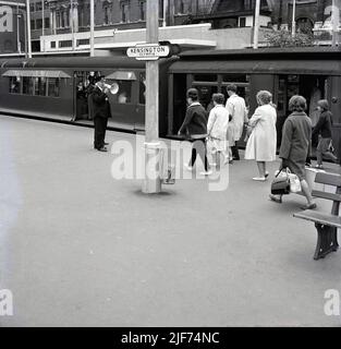 1960s, historique, passagers de train marchant pour monter à bord de la ligne District à la gare de Kensington Olympia, Kensington, Londres, Angleterre, Royaume-Uni. Garde de train en uniforme avec mégaphone sur la plate-forme annonçant le départ. Kensington (Olympia), une station de métro combinée, est desservie par une courte section de la ligne District depuis 1946, initialement dans le cadre du Middle Circle, un service de transport en commun à Londres qui a fonctionné de 1872 à 1905. Le nom de la station vient de son emplacement à Kensington et du centre d'exposition Olympia adjacent et a été ouvert à l'origine en 1844. Banque D'Images