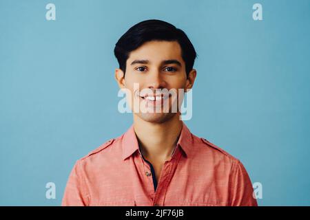 Headshot hispanique latino homme noir cheveux souriant beau jeune adulte portant une chemise rose sur fond bleu regardant la prise de vue en studio Banque D'Images