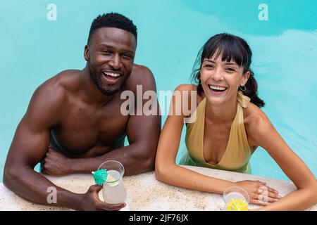 Portrait d'un jeune couple multiracial joyeux prenant des boissons dans la piscine Banque D'Images