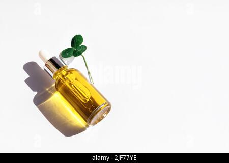 Maquette de bouteille en verre compte-gouttes avec couvercle en métal, huile essentielle et feuille de trèfle curatif sur fond blanc. Sérum, cosmétiques, huile de massage. Lumière naturelle, superbes ombres orange. Copier l'espace Banque D'Images