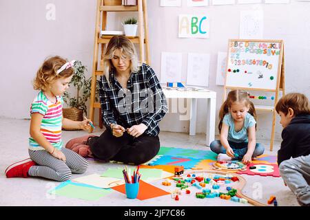 Petits enfants et éducateur plient lego et dessinant avec des crayons et des marqueurs de couleur placés sur le sol dans la salle de jeux. Leçon intéressante pour les jardins d'enfants Banque D'Images