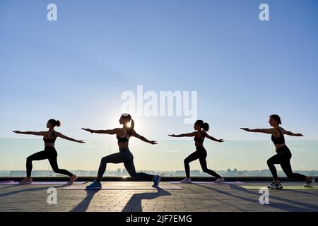 Les silhouettes femelles se posent en guerrier de yoga lors d'un cours de groupe à l'extérieur au lever du soleil. Banque D'Images
