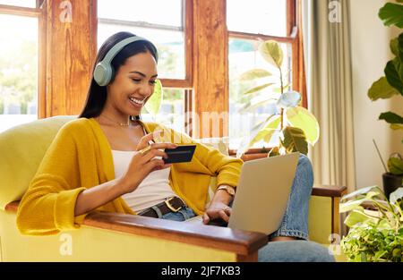 Femme magasiner en ligne tout en tapant sur son ordinateur portable et en tenant sa carte bancaire assise sur une chaise dans un salon lumineux devant une fenêtre. Un jeune Banque D'Images