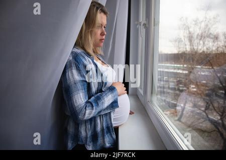 Une jeune femme enceinte vêtue à la maison regarde par la fenêtre Banque D'Images