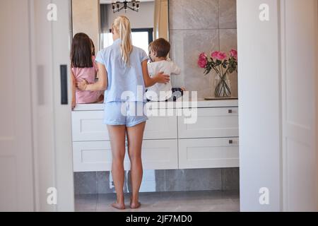 Vue arrière d'une jeune mère caucasienne en pyjama debout avec ses enfants pendant qu'ils se brossent les dents dans la salle de bains à la maison. Petite sœur Banque D'Images