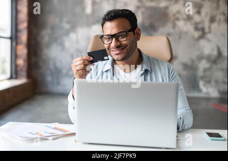 Homme d'affaires indien prospère à lunettes, souriant, assis à l'ordinateur portable, payant sur Internet, acheter en ligne, travaillant à partir de bureau, s tenant une carte de crédit, acheter en ligne, faire des transactions Banque D'Images