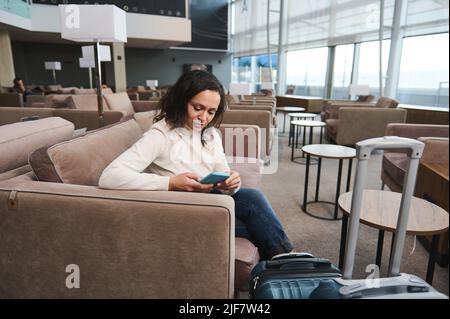 Belle jeune femme du Moyen-Orient en tenue décontractée utilisant le téléphone mobile, la navigation sur les sites Web, la réservation d'hôtel tout en se reposant sur un canapé confortable dans le VI Banque D'Images