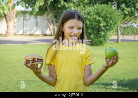 Une adolescente choisit entre un hamburger et une pomme en plein air dans le parc. Banque D'Images
