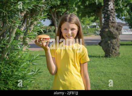 Bonne adolescente mangeant un hamburger dans le parc à l'extérieur. Banque D'Images