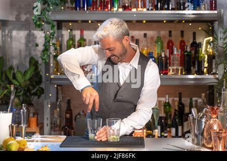 Un barman heureux et charmant prépare un cocktail mojito en boîte de nuit, prépare des boissons expertes sur le comptoir du bar souriant. Banque D'Images