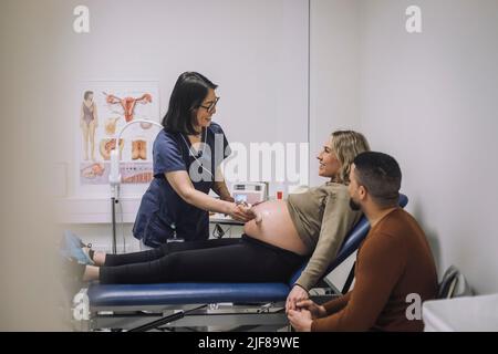 Femme souriante médecin effectuant l'échographie d'une femme enceinte assise sur la table d'examen par un homme à l'hôpital Banque D'Images