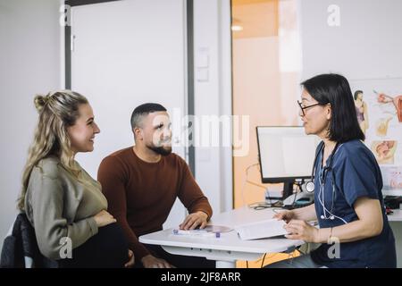 Femme souriante médecin discutant avec une femme enceinte assise par l'homme à un bureau dans la clinique Banque D'Images