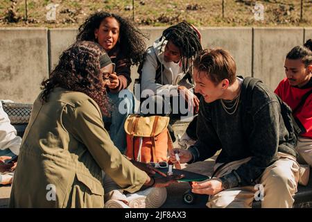 Homme souriant appliquant du vernis à ongles sur la main de la femme tout en étant assis par des amis dans le parc Banque D'Images