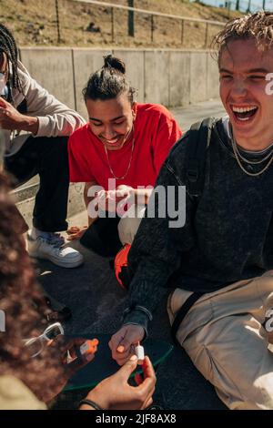 Femme peignant l'ongle d'un homme heureux assis par des amis dans le parc le jour ensoleillé Banque D'Images