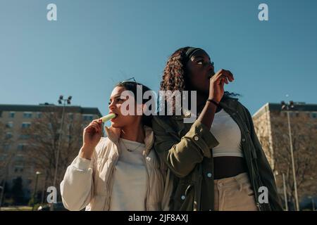 Vue à angle bas des amies qui mangent de la glace tout en regardant loin par temps ensoleillé Banque D'Images