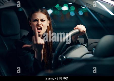 une femme en colère et agressive s'assoit derrière le volant d'une voiture portant une ceinture de sécurité et montre un geste négatif à la caméra hurlant émotionnellement Banque D'Images