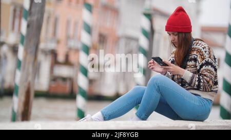 Une jeune femme attrayante s'assoit près du front de mer et regarde inti le téléphone dans les rues de Venise Banque D'Images