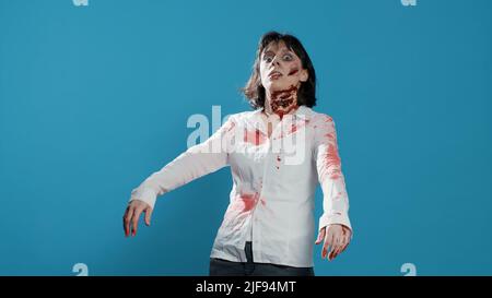 Femme zombie créepy agissant bizarre et sinistre tout en grognant sur fond bleu. Monstre diabolique et inconscient, avec des blessures profondes et sanglantes et des cicatrices qui regardent la caméra. Prise de vue en studio Banque D'Images