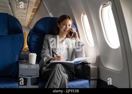 Jeune femme d'affaires asiatique attrayante et prospère parlant au téléphone et travaillant dans la chaise de son avion d'affaires privé Banque D'Images