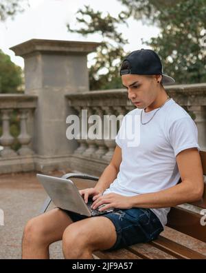 Un cliché vertical d'un gars avec chapeau studiying avec son ordinateur portable sur un banc public Banque D'Images