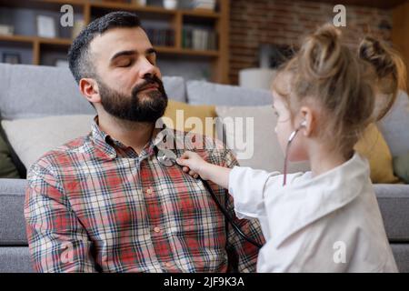 Jolie petite fille jouant docteur avec son père. Drôle d'enfant tient le stéthoscope, à l'écoute de père patient à la maison. Une famille heureuse aime passer du temps Banque D'Images