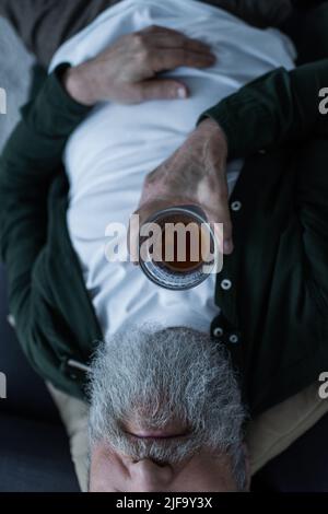 vue rognée d'un homme âgé malheureux avec une barbe allongée sur un canapé et un verre de whisky tout en souffrant de crise Banque D'Images