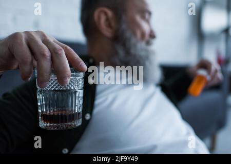 homme âgé flou et malheureux tenant un verre avec le whisky et une bouteille avec des médicaments à la maison Banque D'Images