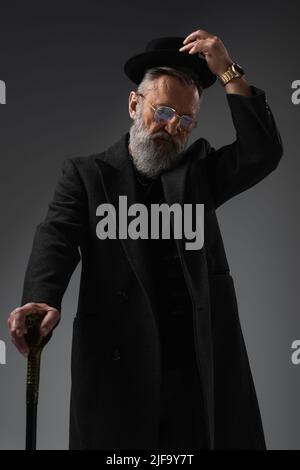 homme senior élégant en manteau élégant portant un chapeau derby et tenant un bâton de marche sur le gris Banque D'Images