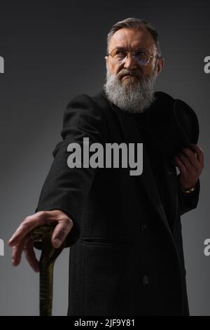 homme senior élégant en manteau élégant tenant un chapeau derby et une canne sur le gris Banque D'Images