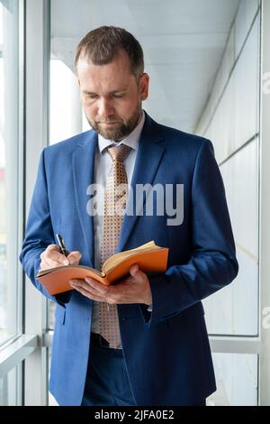 Un homme d'affaires d'âge moyen ciblé en costume écrit des notes dans un journal personnel, planifie une journée de travail, vérifie un planning ou écrit des informations importantes par Banque D'Images