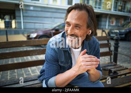 Un beau homme mûr et souriant passe du temps à parler au téléphone ou à écouter de la musique à l'aide d'écouteurs sans fil, assis sur le banc dans une ville urbaine, en appréciant un café portant un Jean bleu. Banque D'Images