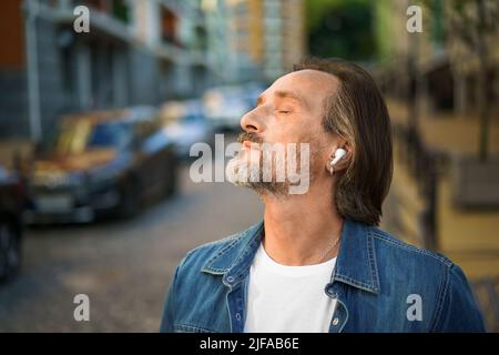 Profiter de la vie écouter de la musique avec les yeux fermés debout à l'extérieur rue de la vieille ville beau homme mûr barbu gris avec des écouteurs sans fil portant une tenue décontractée. Concept de mode de vie de voyage. Banque D'Images