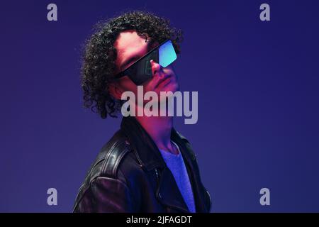 Attrayant homme curly cuir veste porter miroir lunettes regarde la caméra posant isolé sur fond bleu rose. Offre tendance. Immense Banque D'Images