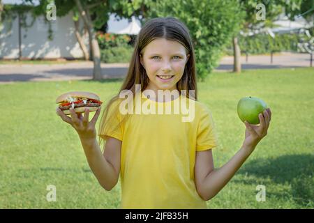 Une adolescente choisit entre un hamburger et une pomme en plein air dans le parc. Banque D'Images