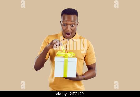 Un homme surpris avec intérêt décompose un cadeau longtemps attendu, isolé sur fond beige clair. Banque D'Images