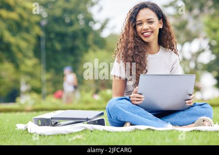 Une jeune femme hispanique travaillant sur son ordinateur portable tout en étant assise à l'extérieur sur un terrain ouvert. Une belle étudiante de race mixte souriant tout en l'utilisant Banque D'Images