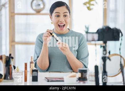 Une belle jeune femme mixte de course se sentant excitée et parlant à la maison tout en l'appareil photo vantant un nouveau produit de maquillage. Un spécialiste de la beauté hispanique stupéfié Banque D'Images