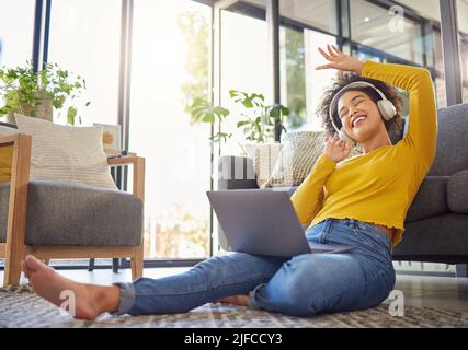 Jeune femme joyeuse de course mixte portant des écouteurs et écoutant de la musique tout en utilisant un ordinateur portable à la maison. Une femme hispanique gaie avec une frino curieuse Banque D'Images