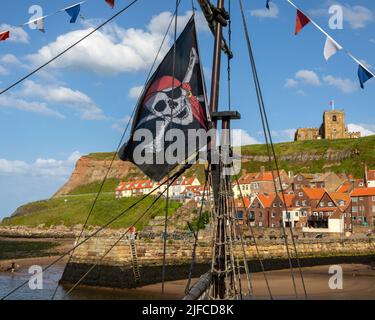 Whitby, Royaume-Uni - 10 juin 2022 : drapeau Jolly Rodger volant sur un bateau pirate dans le port de Whitby dans le nord du Yorkshire, Royaume-Uni. L'église Saint-Marys est visible dans Banque D'Images