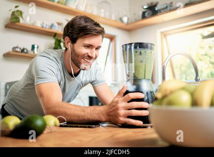 Un jeune homme caucasien en utilisant le blender pour faire smoothie vert sain de détox tout en portant des écouteurs dans la cuisine à la maison. Gars ayant des fruits frais Banque D'Images