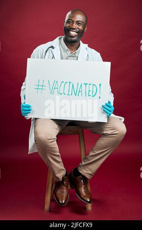 Un médecin afro-américain covid tient et montre une affiche. Portrait complet d'un médecin noir souriant isolé sur fond rouge de studio avec Banque D'Images